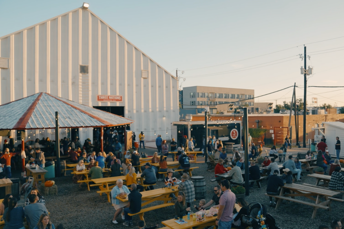 Outdoor patio at Three Nations Brewing with yellow picnic tables, a covered seating area, and people enjoying drinks and food under a clear sky.