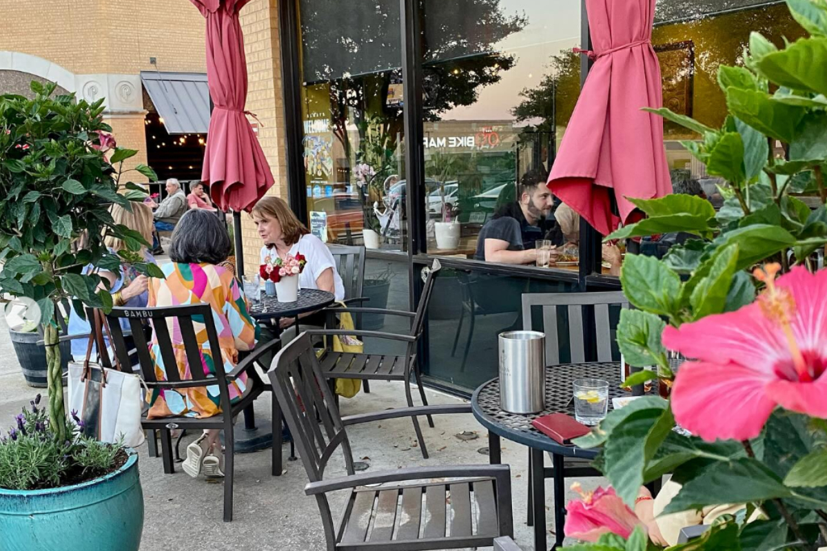 Outdoor patio at Bambu with people dining at small tables, colorful flowers, red umbrellas, and potted plants creating a cozy, inviting atmosphere.