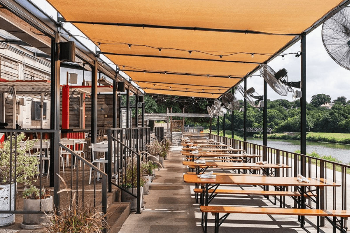 Outdoor patio at HG Sply Co. with shaded wooden picnic tables, ceiling fans, and views of green trees and a river in the background.