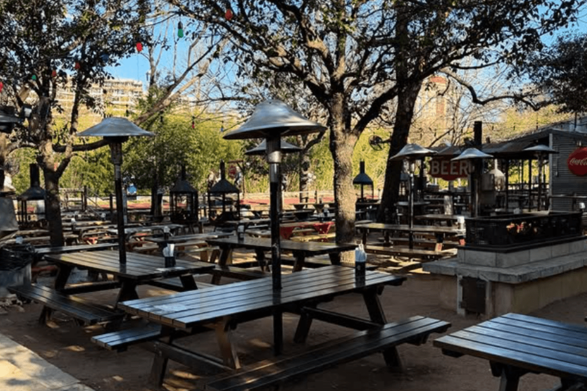 Outdoor beer garden with wooden picnic tables, heaters, string lights, and trees, offering a casual open-air space for dining and drinks.