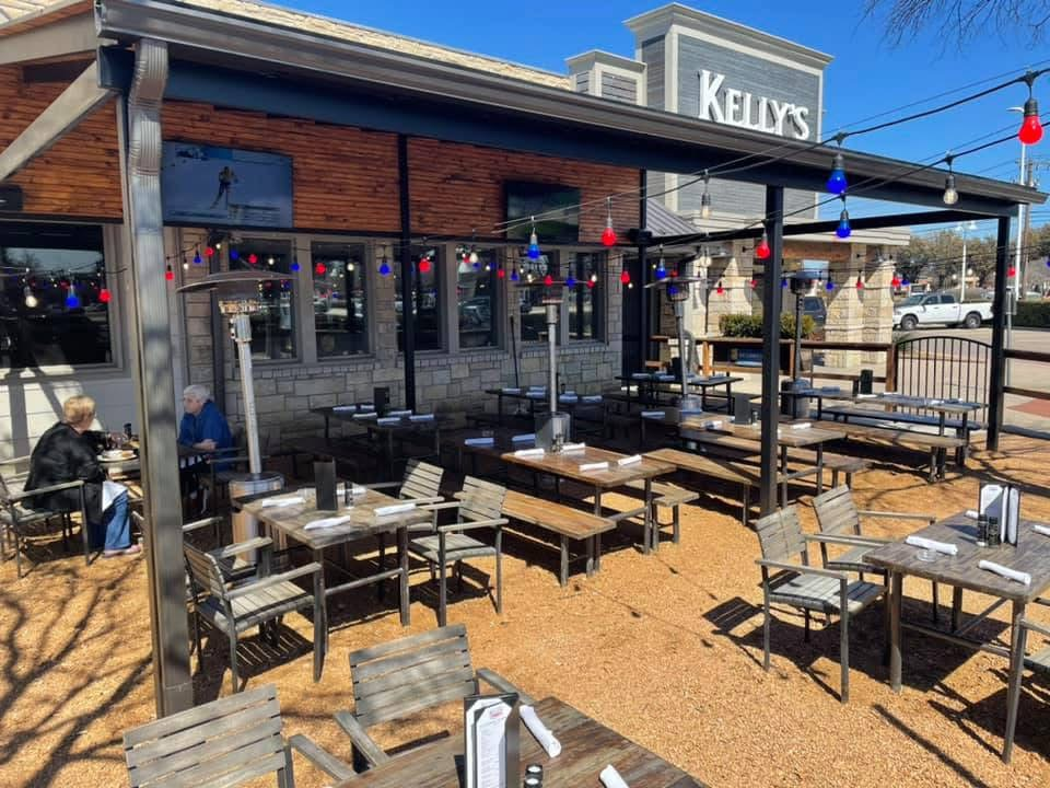 Outdoor patio at Kelly's Craft Tavern with wooden tables, chairs, string lights, and two people dining under a clear blue sky in The Colony.