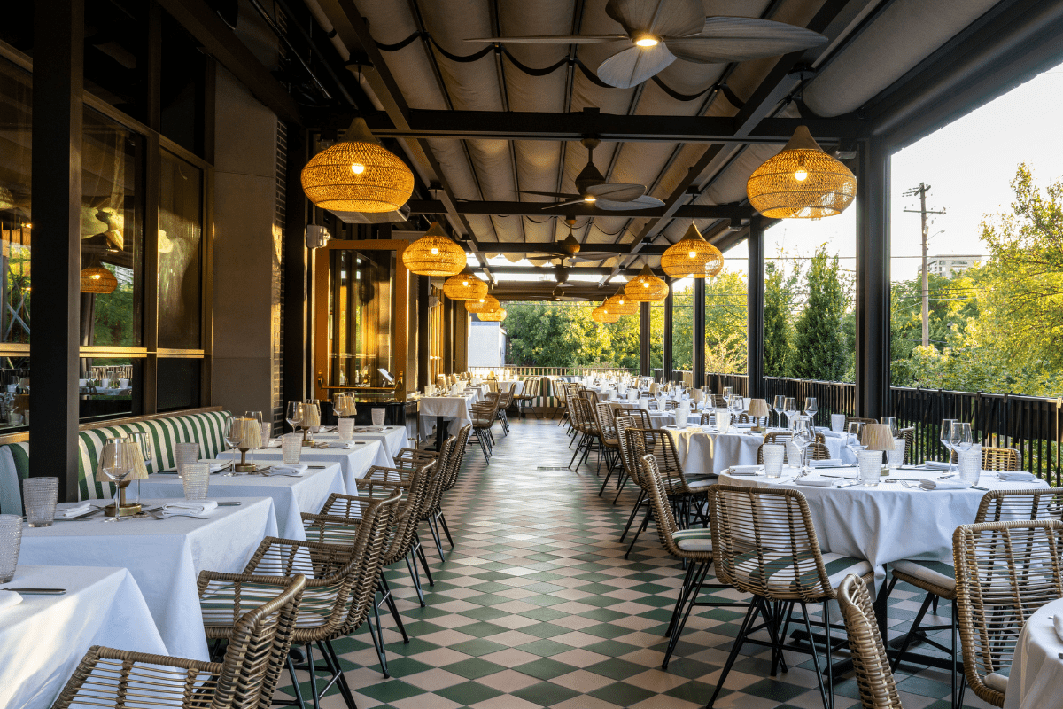 Outdoor patio at Le Passage with elegant tables, white linens, wicker chairs, hanging woven lights, ceiling fans, and a view of trees in the background.