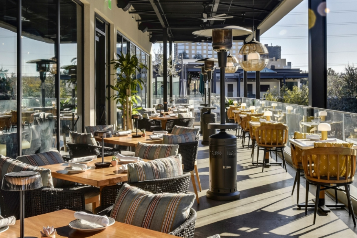 Outdoor patio at Mexican Sugar with wooden tables, modern wicker and cushioned chairs, hanging lights, patio heaters, and glass railing with city views.