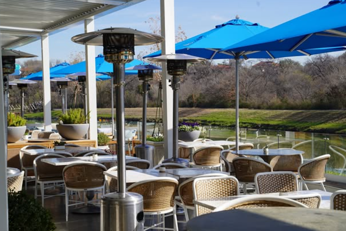 Quince Riverside outdoor dining with wicker chairs, white tables, blue umbrellas, and patio heaters, overlooking a peaceful, tree-lined river view.